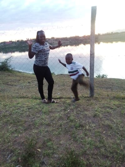 This image depicts a joyful outdoor scene featuring a woman and a young boy in Central Kasipul ward, Kenya, likely captured during the late afternoon or early evening. The light is soft and diffused, indicating either sunset or a slightly overcast sky.
In the foreground, a dark-skinned woman with long, dark braided hair is positioned on the left. She smiles broadly at the camera, raising both hands in a friendly wave. She wears a light-colored, short-sleeved t-shirt featuring repeating text designs, including the word "MONTE" and other jumbled letters, paired with dark leggings and light-colored sandals. She leans slightly against a tall, light-colored fence post that marks the right edge of the frame.
To her right, a young dark-skinned boy is caught in mid-action, appearing to run or skip. His left leg is raised, and his right arm is extended, conveying a sense of energy and playfulness. He wears a white short-sleeved t-shirt with horizontal green, white, and red stripes across the chest, and brown shorts or trousers. His face is slightly blurred due to motion.
Both individuals stand on a grassy and earthy bank. Behind them, a chain-link fence stretches horizontally, acting as a boundary to a large, tranquil body of water that dominates the background. The water beautifully reflects the soft light of the sky above, creating a serene backdrop. In the far distance, across the water, indistinct hills or land formations are visible. The overall scene conveys a happy and energetic moment in a peaceful natural setting.