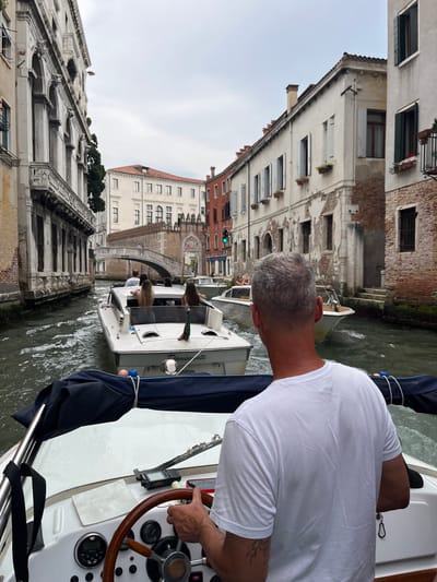 A medium-angle shot depicts a man piloting a boat through a Venetian canal, likely the Grand Canal, in Venice, Italy. The scene is viewed from behind the pilot, who is Caucasian, with short gray hair, and wearing a white T-shirt. He is holding the wooden steering wheel of the boat, which features various gauges and equipment on its dashboard. His right arm is visible, showing a tattoo on his upper arm.
In the background, a canal is bordered by old Venetian buildings, constructed from light-colored stone or brick with a variety of window designs. On the opposite side of the canal, a similar boat is traveling in the same direction, carrying several passengers. Further back, the arched Rialto Bridge is visible, a prominent landmark in Venice. A green traffic light is seen near the bridge. The sky above appears overcast. The water in the canal shows movement from the boats.