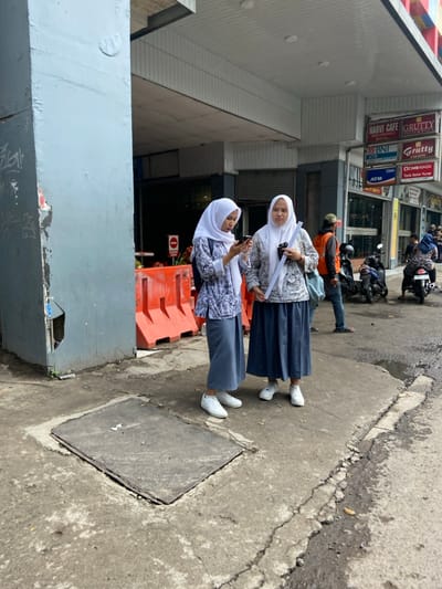 Two women, both wearing white headscarves and long grey skirts, are standing on the sidewalk in front of a building in Bandung, Indonesia. The women are both looking at their phones and are standing next to each other. There are orange construction barriers to their left and a large metal grate in the foreground. There are signs in the background for a cafe, ATM, and a bank. A man wearing a blue shirt and a red cap is walking in the background. There are parked motorcycles behind him. The sidewalk is grey and uneven, and there are cracks running through it. The building in the background is white with a gray roof. There is a sign that reads "Harvi Cafe." The image is taken from a slightly elevated perspective. The location is in the city of Bandung, Indonesia, which is located at the coordinates -6.952, 107.593.