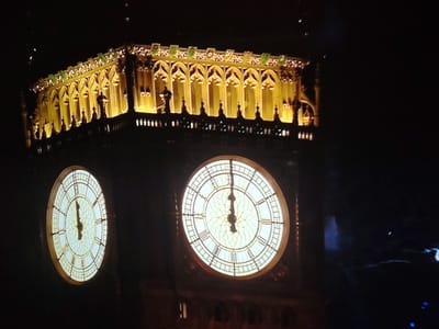 This image captures a night view of the Elizabeth Tower, commonly known as Big Ben, in London, United Kingdom. The iconic clock tower is illuminated, with its large clock faces prominently displayed. The top section of the tower, featuring gothic architectural elements and ornamental details, is also brightly lit with a warm yellow light. One clock face is larger and more centrally positioned in the frame, showing the time as approximately 12:00. A second, smaller clock face is visible to the left, also appearing to indicate 12:00. The background is dark, indicating nighttime, with some faint lights or reflections in the lower right portion of the image. There are no people or active interactions visible in the scene.
