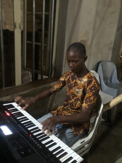 An image, correctly oriented, depicts a young man playing a black digital keyboard in an indoor setting. He is positioned in front of the instrument, with both hands on the keys, seemingly engaged in playing music. The man has dark skin and short hair, and is wearing a short-sleeved shirt featuring a distinctive brown, gold, and black geometric pattern, possibly an Ankara fabric, along with light blue trousers.
The keyboard displays small red indicator lights and a glowing screen, suggesting it is powered on. The overall lighting in the room is dim. In the background, to the left of the man, a structure composed of horizontal metal bars is visible, possibly part of a railing or window grill. Behind him, a grey plastic chair is partially visible, along with other indistinct elements of what appears to be a utilitarian or minimally furnished room. The man's expression is one of focus on the activity. No discernible text is visible within the image. The scene is likely set in Ukwu-Abwa, Nigeria.