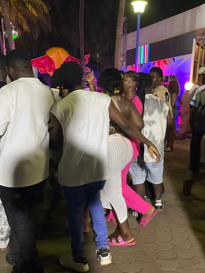The image depicts a nighttime outdoor social event in Accra, Ghana, characterized by artificial lighting and a lively crowd. In the foreground, multiple individuals of varying skin tones are observed. From left to right: a male figure with short dark hair, seen from behind, wears a white short-sleeved t-shirt and dark pants. Next to him, another male figure with dark, possibly braided hair, also seen from behind, wears a white sleeveless top and blue jeans, with white sneakers. His right arm is raised. Adjacent to him, a female figure with dark hair pulled back wears a light brown halter-neck top, a white long mesh-knit skirt over bright pink pants or leggings, and pink slide sandals. Her left arm is extended towards another female figure in a bright pink top, whose face is partially obscured. Further right, a male figure is visible from the back in a white tie-dye sleeveless shirt and light blue denim shorts. Several other individuals are visible in the mid-ground and background, some appearing to socialize or dance.

The environment includes several tall palm trees silhouetted against the dark sky. A prominent inflatable Red Bull logo (red bull, yellow sun disc) is visible in the left background, indicating brand sponsorship. To the right, an outdoor stage or backdrop structure features vertical multi-colored panels, resembling a test pattern, and is illuminated by purple and blue lighting. An overhead street lamp with a yellow glow is positioned in the upper right. The ground is paved with interlocking brick patterns. The scene suggests an active, festive gathering.