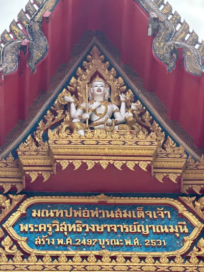 The image shows the upper facade of a temple in Chalong, Thailand. A gilded, multi-armed statue of a deity is prominently featured at the apex of the structure, adorned with intricate gold ornamentation. Below the statue, blue tiles form a backdrop for Thai script inscribed within a gold border. The script, transliterated, reads: "Manthop Pho Than Somdet Chao Phra Khru Wisutthi Rangsithan Muni." Below this, it states: "Khrang, Ph.S. 2497, B.E. 2531." The architecture displays vibrant red walls and ornate, serpent-like decorative elements, also covered in gold and mosaic tiles. The overall scene is a detailed view of religious architecture and statuary.