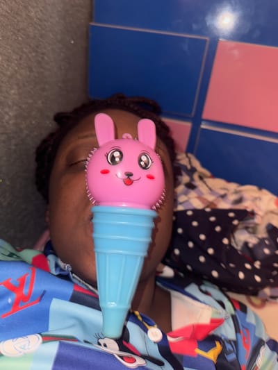 A close-up, tilted shot reveals a child lying down, with a pink and blue rabbit-shaped object positioned near their face. The child's hair is dark and braided. The child is wearing a patterned shirt with various colors and designs, including blue, red, and white elements. Part of another patterned fabric, predominantly dark blue with white polka dots, is also visible in the background. The floor is mostly gray and reflective, with blue and pink square tiles visible along one edge of the frame. The location, Sabon Tasha, Nigeria, is not evident in the image.