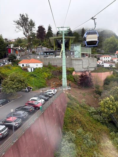 The image shows a view from a cable car in Funchal, Portugal, looking down at the ground below. A blue and white cable car is visible in the upper left of the frame. Below the cable car infrastructure, buildings and trees are visible on a hillside. There is a parking lot on the lower right, filled with several cars, including red, white, and grey vehicles. The ground and some parts of the view are affected by mist or haze.