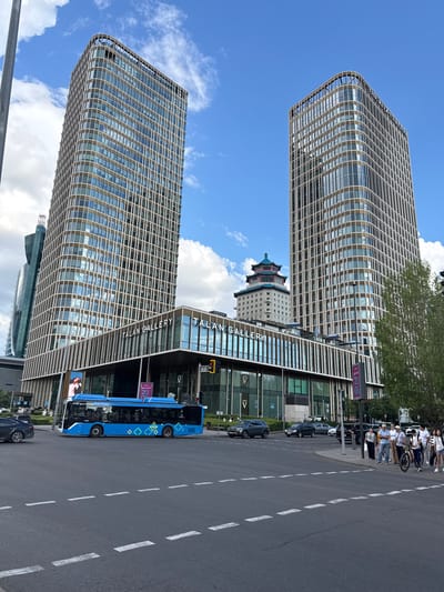 The image depicts a street scene in Astana, Kazakhstan, on a bright, sunny day with scattered clouds. Dominating the view are two modern, multi-story buildings with distinctive geometric facades featuring a lattice-like pattern of light-colored beams and glass windows. In the background, a traditional building with a tiered, pagoda-style roof is visible, suggesting a blend of architectural styles.

In the foreground, a blue bus with a diamond-patterned design is parked along the side of the road. Several cars are also parked nearby, and a few people are visible walking on the sidewalk and standing on a crosswalk. Some individuals are also seen in the background, near the buildings, and a group of people are gathered further down the street, some of whom appear to be looking at something or engaged in conversation.

Visible text includes "TALAN GALLERY" on the facade of one of the buildings. Another sign on the bus reads "AYEL GAZDERİNA". The overall impression is a contemporary urban environment with hints of historical architecture, under clear, daytime skies. The mood appears to be calm and ordinary, depicting a typical day in the city.