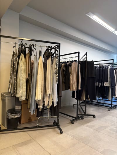 The image depicts an interior scene, likely a clothing store or showroom, in Milan, Italy. Multiple black metal clothing racks are set up on a light-colored tiled floor. The racks are laden with various garments, primarily scarves and formal wear such as suits and jackets. The color palette is subdued, dominated by neutral tones like grey, beige, and black, with some items featuring subtle patterns. In the foreground, on the left side, a tall silver waste bin and a smaller black trash can are visible. Behind the clothing racks, a light grey wall serves as the backdrop, with a long fluorescent light fixture visible on the ceiling. The overall impression is one of an organized retail or display space, emphasizing a sophisticated and minimalist aesthetic.
