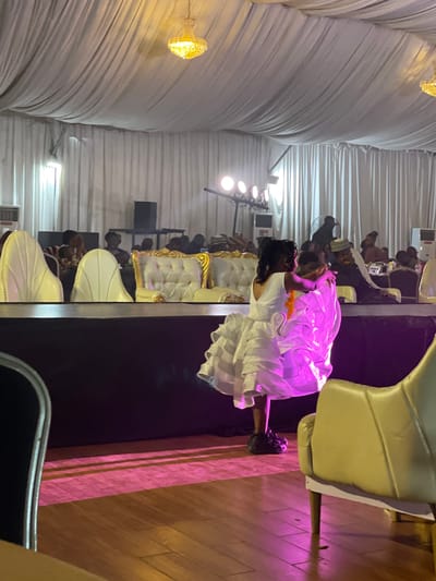An indoor event is underway at the Barakin Lerawi, Nigeria event hall, characterized by draped white fabric ceilings adorned with golden chandeliers and lined with white pleated drapes. A raised stage area is set up with plush, white, tufted seating with gold trim. A young girl in a voluminous white dress and dark shoes is the central focus, her back to the camera, appearing to embrace or hold onto something or someone not fully visible. The floor is polished wood, illuminated by pink stage lighting creating striped patterns. In the background, guests are seated at tables and in chairs, with some individuals visible behind the stage, including a man wearing a cap and a person at a table labeled "TABLE 4". Air conditioning units are visible on the sides.