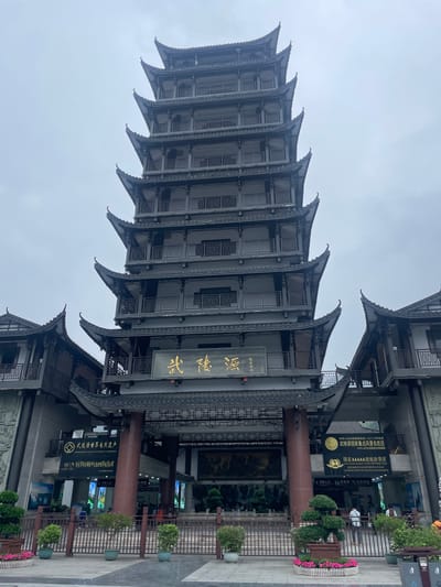 The image shows a tall, traditional Chinese building in Wulingyuan, China. The building has multiple levels, each with a curved roof. There is a sign with Chinese characters on it. To the right of the building, there is a sign indicating that it is a National AAAAA tourist attraction and also indicating the Wulingyuan World Natural Heritage site. Additional signs advertise that it is a key scenic area. The sky is overcast. A sign indicates entrance for line A, as well as information about toilets and boiled water. There are small potted plants surrounding the base of the building.
