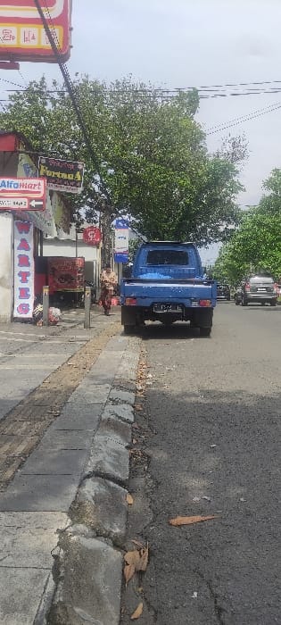 The image shows a street scene in Bogor, Indonesia, at the coordinates -6.584 latitude and 106.818 longitude. A blue pickup truck is parked on the street, facing away from the camera. The truck has a license plate that reads "D 1214". There are a number of power lines running overhead, with some of them broken. The road is paved and there is a sidewalk to the left of the road. The sidewalk is made of concrete and has some cracks in it. There are a few fallen leaves on the road. There are several trees in the background and a person in the distance walking towards the truck. The person is wearing a yellow shirt. On the left side of the image, there is a building with a sign that reads "Alfamart." The building has several signs, including one that reads "Fortuna 4". The image was taken from a low angle, looking towards the street.