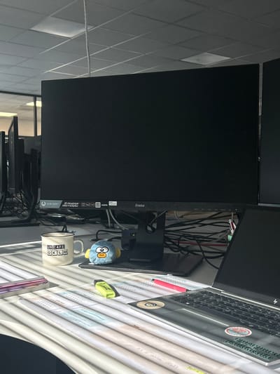 The image displays an indoor office workstation located in Reichstett, France. The scene is illuminated by strong natural light entering from the left, casting distinct horizontal shadow stripes from venetian blinds across the desk and its contents. A large, black Iiyama G-Master computer monitor, featuring a dark screen, stands centrally on a black base. To the left of the monitor, a white ceramic mug with "UN CAFE OKLM" printed in black text and a small, light blue plush toy with a yellow beak are positioned. To the right, an open silver HP laptop with a dark screen is visible, featuring multiple stickers on its lid. The white desk surface in the foreground holds several sheets of paper with handwritten notes, a yellow-green highlighter pen, and a pink pen. Behind the central monitor, a dense tangle of black cables is observable. In the background, other workstations with multiple dark computer monitors are partially visible, and the ceiling features square acoustic tiles with integrated lighting fixtures. No individuals are present in the image, and no active interactions are depicted.