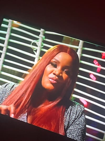 A medium shot captures a Black woman with long, auburn hair and a sequined silver top. She is looking towards the left side of the frame, her mouth slightly open as if speaking. Her left hand is visible in the lower left corner, gesturing forward. Behind her, a slatted white structure with green accents stretches across the background, punctuated by bright pink, oval-shaped lights. The lighting suggests an indoor setting, possibly a studio or performance space.