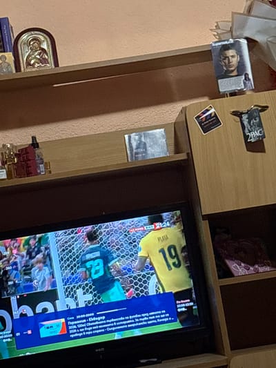 The image depicts an indoor scene, likely a living room or bedroom, with a television set as the central focus. The TV displays a soccer match, indicated by the players in uniform, a net, and visible text overlays on the screen providing match information. The text suggests the game is between Germany and Ecuador, part of the 2026 World Cup, and indicates a time frame of 20:00-22:00.

On a shelf above the TV, several objects are visible. These include a framed picture or card with a portrait of a person, possibly a celebrity, and another item with "2PAC" written on it, suggesting a connection to the rapper Tupac Shakur. There are also smaller stickers or cards on the shelf.

The overall setting appears to be a personal space, with decorative items and personal memorabilia suggesting a lived-in environment. The lighting is dim, indicating it might be evening or nighttime, consistent with the TV broadcast schedule. There are no clear indications of weather conditions. The scene conveys a casual, home-based activity of watching television.