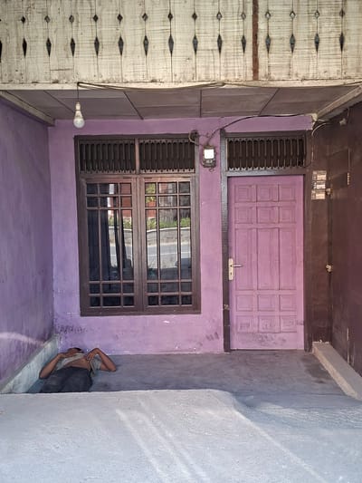 A man is lying on his back, asleep or resting, on a concrete floor outside a purple-painted building. He is wearing a light-colored t-shirt and dark pants. His head is tilted back, and his arms are resting beside him. The building has a wooden lattice structure above a light fixture and a tiled ceiling. To the left of the man is a window with dark brown wooden frames and bars. To his right is a purple door with paneled sections and a silver doorknob. Next to the door is a dark brown wooden door frame. The ground is concrete, with a lighter colored concrete barrier in the foreground. The image appears to be taken during the day, with sunlight casting shadows on the ground. The location is Bireuenmeunasahcapa, Indonesia.