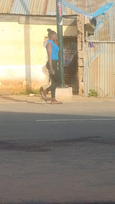 In a medium shot, a young Black woman walks along a paved road in Tempure, Nigeria. She is wearing a bright blue sleeveless top and dark leggings with white accents. Her hair is pulled up in a bun. She is walking from left to right across the frame, and her body is angled slightly away from the camera. She wears white flip-flops.
The setting appears to be an urban or semi-urban area. Behind the woman, there is a building with a light yellow facade and a corrugated metal roof. Part of the building is constructed with rough stone. A black metal pole with some graffiti stands in front of the building, and a satellite dish is mounted on the roof. The ground in front of the building is dirt with some sparse vegetation. The road in the foreground is asphalt and shows signs of wear. The lighting suggests it is daytime, likely morning or afternoon, with distinct shadows cast by the building and the woman. The overall mood is casual and everyday. There is some text visible on the woman's leggings, appearing as two concentric circles with white outlines. There is also a faint "A" logo visible on the pole.