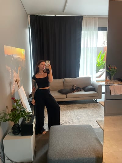 The image captures a full-length mirror selfie taken by a woman inside a modern, well-lit room. The woman is standing and wearing a black crop top and black wide-leg pants. She has a pearl necklace on and is holding her phone in her right hand, taking the picture. Her left arm is extended downwards, and she has tattoos on her left arm. The background reveals a living area with a light-colored sofa where a small dog is resting. Behind the sofa, sheer white curtains are drawn back, revealing a glimpse of an outdoor area with lounge chairs and lush greenery. To the left of the woman, a projector displays a scenic image of mountains and a sunset onto the wall. Beside the projector, an easel holds a large white canvas. In the foreground, a white cube-shaped cabinet holds a vase with flowers and a black and gold speaker. The room's flooring appears to be a combination of a patterned rug and tiled surfaces. The style of the room suggests a contemporary interior design, possibly in an urban setting. The mention of Kediri, Indonesia, is not visually supported by any specific landmarks or cultural indicators within the image itself.