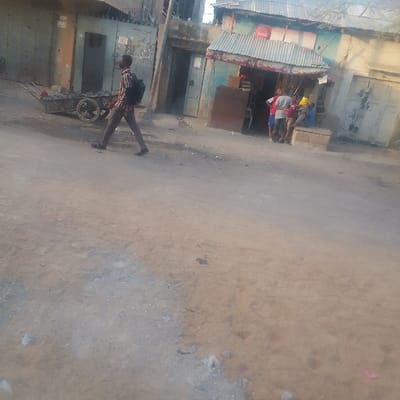 A man with a backpack walks along a dusty street, pulling a cart filled with what appears to be rocks. Across the street, a small store with a corrugated metal roof and a Coca-Cola sign is visible. A few people are congregated outside the store. Buildings of modest construction surround the area. The scene seems to depict a street-side setting in a developing or rural area.
