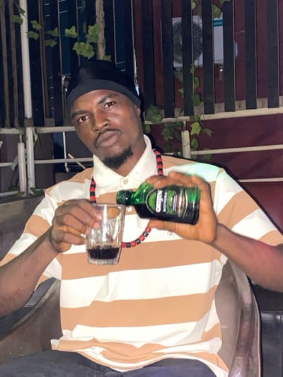 A man in a striped shirt pours a bottle of “Origin” alcohol into a glass. He wears a black head covering and a necklace made of small red and black beads. The background features a partially visible structure with horizontal slats and artificial ivy. The lighting suggests an indoor or nighttime setting.