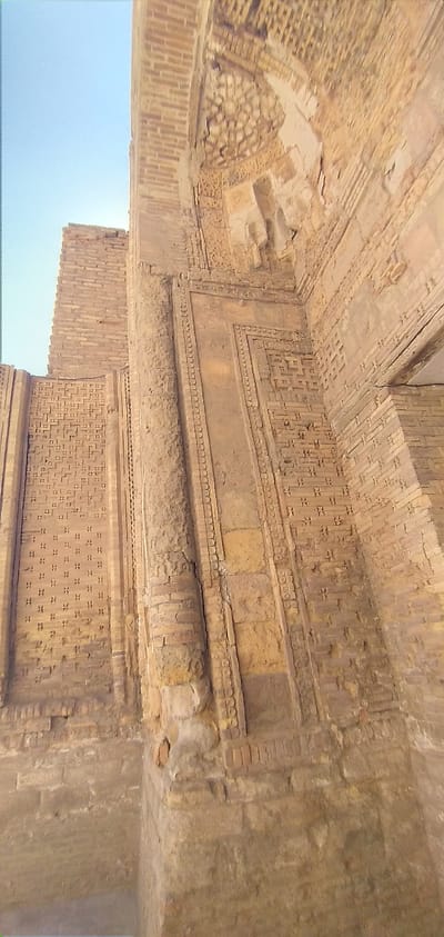 This image shows the Kalon Minaret in Bukhara, Uzbekistan. The minaret is a tall, brick tower with intricate geometric carvings and patterns. The lower part of the tower is visible, with a weathered, rough texture from age. The sky is a clear, pale blue. The image is taken from a low angle, looking up at the minaret. The focus is on the detailed brickwork and architectural features of the minaret.