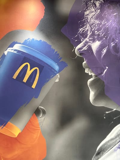 The image is a close-up, abstract photograph featuring a blue McDonald's cup with a golden "M" logo prominently displayed. The cup is held by a hand, partially visible on the left side of the frame, against a background that blends abstract orange and grey shapes. Overlaying the scene, on the right, is a purple-tinted profile of a person's face, emphasizing their nose, mouth, and curly hair. The overall aesthetic is artistic and fragmented, with a focus on color and texture. There is no discernible location in the city of Derry/Londonderry, United Kingdom, visible in this image.