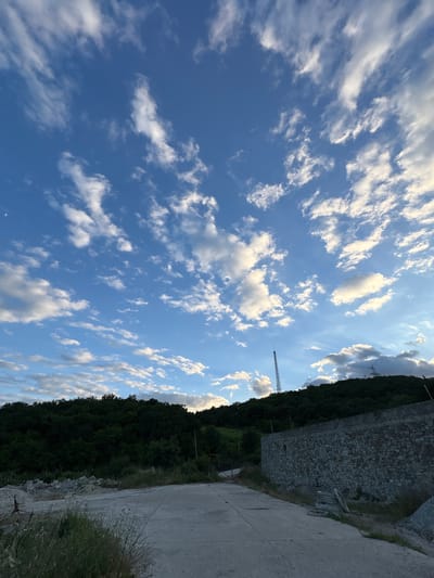 The image captures an outdoor scene with a cloudy blue sky dominating the upper portion. A crescent moon is visible in the far upper left corner. Below the sky is a dark, tree-covered hillside. In the foreground, there is a concrete road or pathway leading into the scene from the bottom. To the right of the pathway, a stone wall is constructed. The environment appears somewhat rural or undeveloped, with some piles of rocks and gravel visible on the left side of the pathway. A tall communication tower is visible on the left side of the hillside, and another, larger tower with power lines is visible on the right side of the hillside. The lighting suggests it is either early morning or late afternoon.