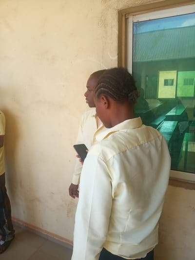 Two individuals, possibly men, are captured standing next to a cream-colored wall and a tinted window, likely in an indoor or covered outdoor setting. The person in the foreground is viewed from behind, wearing a light yellow long-sleeved collared shirt. Their dark hair is styled in braids pulled back. They are holding a black smartphone in their right hand.

To the left, a second person is partially visible in profile, also wearing a similar light yellow long-sleeved shirt. A third person's lower leg and foot, wearing dark sandals, are seen on the far left edge of the frame.

The window features green and blue tinted glass, reflecting what appears to be other nearby buildings with yellow and green exteriors and visible windows, along with a glimpse of a light blue sky or structure. The wall behind the individuals is a plain, light cream color, showing some signs of wear and dirt. A light-colored tiled floor is visible at the bottom.

The scene suggests a casual daytime gathering or waiting period in Nsugbe, Nigeria, with the individuals appearing neutral in mood, possibly conversing or awaiting an event. No text is visible in the image.