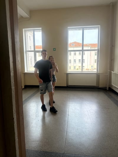 Two young individuals, a male and a female, stand in an empty, bright room with large windows. The male, in the foreground, wears a black t-shirt and ripped denim shorts. The female, slightly behind and to his right, wears a light-colored tank top and denim shorts, and appears to be looking towards the male. The room has a concrete floor and beige walls. Two large windows, divided into panes, reveal buildings and a cloudy sky outside. White radiators are visible beneath the windows. A green directional arrow sign is mounted on the wall between the windows. The overall impression is of an interior space, possibly part of a school or institutional building. This image was taken inside the former school building of the Vasil Levski National Military University in Shumen, Bulgaria.