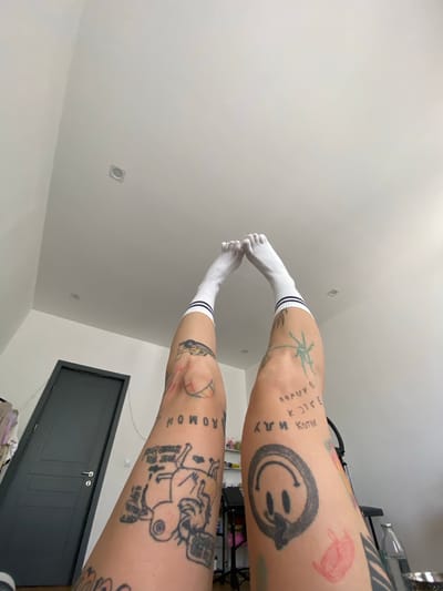 This is a first-person, low-angle, indoor shot of a person's legs, which are raised and bent at the knees, reaching towards a white ceiling with recessed lighting. The person is wearing white socks with two dark stripes near the top. Both legs are heavily tattooed with various designs, including a smiley face, a basketball, a cartoon character, and text in what appears to be Cyrillic script. The background features a dark grey door with a silver handle on the left and a shelf with bottles and a black chair on the right. The overall environment appears to be a residential room. The specific location of Tbilisi, Georgia is not determinable from the visual information provided in the image.