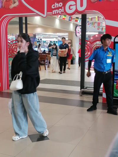 A young East Asian woman with long dark hair walks left to right across a tiled indoor walkway in what appears to be a shopping mall. She is wearing a black hooded sweatshirt, loose light blue jeans, and white slip-on shoes. She is carrying a white handbag on her left shoulder and a clear plastic bag in her left hand. Her right hand is near her face.
To her right, further in the background, a man in a blue uniform with black pants and a black belt walks right to left. He has short dark hair and is wearing black shoes. He appears to be a security guard.
Behind the woman, a man in a dark blue t-shirt and black pants is walking right to left, carrying a cardboard box. A woman in a red dress and a light gray cardigan walks behind him.
The background shows a red and white curved awning with text in what appears to be Vietnamese. Flags are hung across the top of the awning. The floor is tiled with light beige and darker beige square tiles, arranged in a diamond pattern. The lighting is bright and diffused.