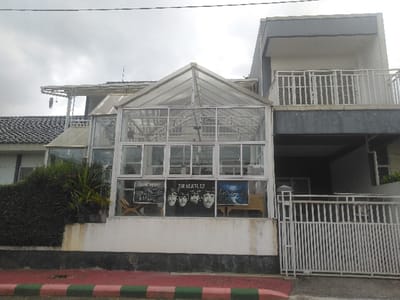 The image displays a multi-structure property situated in Kladno, Czechia, under an overcast sky. The central element is a two-story, predominantly glass-paneled structure with white framing, reminiscent of a conservatory or sunroom. Within its lower level, four distinct black and white images of The Beatles are visible, accompanied by the text "THE BEATLES." To the immediate right, a modern-style building features a grey and white facade, with a second-floor balcony enclosed by white railings. The ground level below this balcony appears to be a recessed entrance or garage area. To the left of the glass structure, a segment of an older building with a dark tiled roof is partially visible, obscured by a dense green hedge. The foreground features a low white wall with a dark base, continuing as a white metal gate providing property access. A street curb, painted with alternating red and green stripes, marks the edge of an asphalt surface at the bottom of the frame. Thin utility lines are visible against the sky at the top. No individuals are present in the scene.