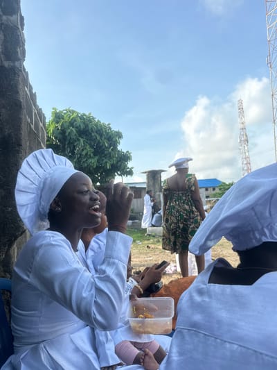 The image depicts an outdoor gathering in Badagry, Nigeria, under a bright, partly cloudy sky.
In the left foreground, a young woman with dark skin is prominently featured from the chest up, positioned against a gray concrete block wall. She wears a white, pleated, chef-style hat and a long-sleeved white garment. Her mouth is open, indicating she is speaking or singing, and her left hand is raised to her mouth, while her right hand is partially visible further back. A small gold earring is visible on her left earlobe. Behind her, another individual in similar white attire is partially visible.
In the immediate lower foreground, parts of two bare feet, one with a pink bandage or fabric wrapping, are visible next to a clear plastic container holding a yellow-orange granular food, likely rice. Hands are partially visible holding both the container and a dark-colored mobile phone.
In the mid-ground, a woman with her back to the camera stands wearing a sleeveless, patterned dress in shades of green, brown, and black, along with a white hat similar to the foreground subject's. To her left, another person in a long white robe is partially visible near a weathered concrete structure. The ground in the mid-ground is unpaved, appearing as dirt or sparse grass.
The background features several structures. To the far left, a dense green tree provides foliage. Towards the center-right, two communication towers, one larger and more prominent than the other, extend into the sky. Scattered buildings with light-colored walls and one with a blue corrugated roof are visible between the figures and the towers. The overall scene is brightly lit by natural sunlight, casting minimal shadows.