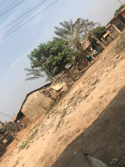 The scene depicts an outdoor, semi-developed area in Ago-Abo, Nigeria, under a clear, hazy light blue sky with power lines visible in the upper left quadrant. The ground consists of uneven, light brown dirt with sparse patches of green vegetation and scattered debris. In the mid-ground left, a structure constructed from grey concrete blocks is visible, adjacent to a makeshift stall or storage area covered with a light-colored fabric tarp and supported by wooden frames. A prominent green-leafed tree stands centrally, with additional trees, including one with elongated leaves, further back. In the background right, residential structures are present, some with unpainted block walls and others with sections of reddish-brown coloring. Laundry is hanging on lines near these structures, and two to three indistinct figures are discernible in the far background. A diagonal shadow extends across the lower right portion of the ground.