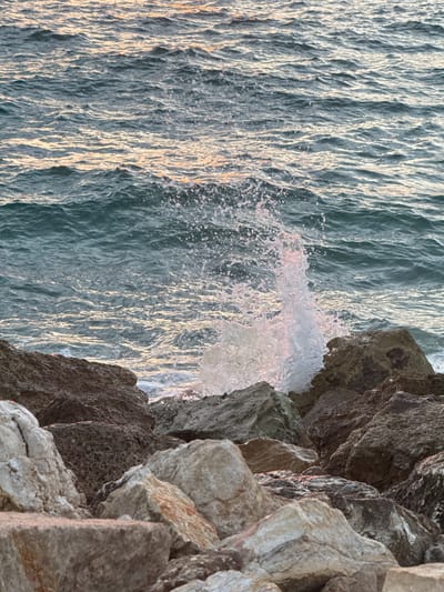 This media file captures a close-up view of a wave crashing against a rocky coastline in Tel-Aviv, Israel. The scene is dominated by the texture and movement of the water and the rough, angular surfaces of the rocks.
The water occupies the left side of the frame, appearing as a dynamic expanse of rippling lines and crests. Sunlight reflects off the surface, creating streaks of bright white and golden light that contrast with the deeper blues and greens of the water. In the center, a dynamic splash of water is frozen in mid-air, showcasing the force of the wave. Tiny droplets and foam are visible, hinting at the energy of the impact. The water appears slightly opaque due to the light and the suspended particles.
On the right side of the frame, large, irregular rocks form a rugged barrier. Their surfaces are varied, with some appearing smooth and worn, while others are rough and jagged. The rocks are a palette of grays, browns, and hints of warmer tones, suggesting mineral content or the effect of light. The close perspective emphasizes the texture and dimensionality of the rock formations.
The activity depicted is the natural event of a wave interacting with the shore. The time of day appears to be either sunrise or sunset, as indicated by the warm, low-angle sunlight illuminating the water and highlighting the textures of both the water and the rocks. The weather seems clear, allowing for strong reflections on the water. No people or man-made objects are visible, focusing the attention entirely on the elemental forces at play. The scene evokes a sense of raw natural power and the ever-changing beauty of the coastline. There is no visible text in the image.
