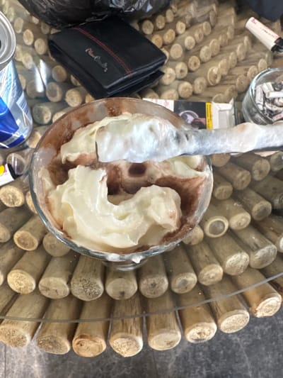 A high-angle shot reveals a glass table with a unique surface comprised of numerous small, cylindrical wooden dowels arranged tightly together. At the center of the frame is a partially filled glass containing a layered dessert, seemingly a chocolate-based beverage topped with whipped cream or a similar dairy product. A partially submerged straw is visible within the glass. To the left of the glass, a black leather wallet with red stitching detail sits, along with a discarded beverage can. In the background, elements such as a lighter, a glass ashtray with cigarette butts, and part of a package/box are visible. The location is unspecified, and there is no indication that it is Yambol, Bulgaria. No people are visible.