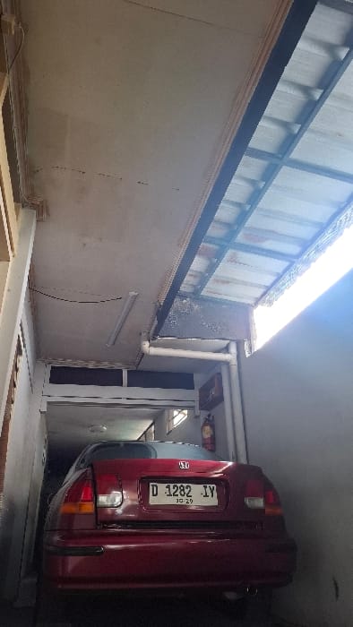 The image shows the rear of a red Honda Civic parked in a narrow, enclosed space. The car has a license plate that reads "D 1282 - IV" and is obscured by the wall. There is a white ceiling above, partially visible, suggesting the vehicle is parked under a carport or a covered area. The location, based on the coordinates, is Bandung, Indonesia, with the scene likely occurring in a residential neighborhood or a private garage. The environment appears to be a typical urban setting with a mix of concrete and metal elements. There are no people in the scene.