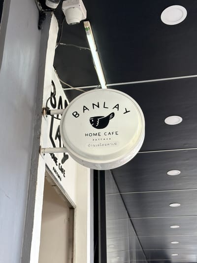 The image shows a circular white sign for "BANLAY HOME CAFE PATTAYA". The sign features a black graphic of a bird and Thai text. The sign is mounted on the exterior wall of a building. Above the sign, a security camera and a fluorescent light fixture are visible. The building appears to be located in an urban environment, possibly a commercial area, given the ceiling with recessed lighting and the presence of a doorway to the right of the sign. The wall to the left of the sign is painted white. The overall scene suggests an establishment in Pattaya City, Thailand.