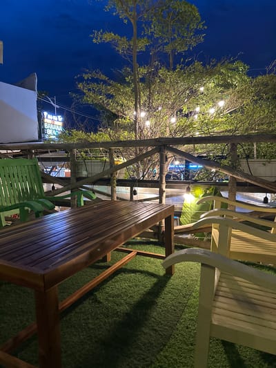 The image depicts an outdoor seating area at night, likely a restaurant or cafe, in Phu Thuy Ward, Vietnam. A wooden table sits on an artificial turf surface, flanked by several chairs. One prominent green wooden chair is on the left, with two lighter-colored wooden chairs on the right. In the background, a wooden railing separates the seating area from a densely vegetated space illuminated by string lights. A neon sign is visible in the upper left, advertising "FOOD & DRINK BANANA BEACH". The sky is a deep blue, indicating it's evening or night. The scene is quiet and still, with no people present.