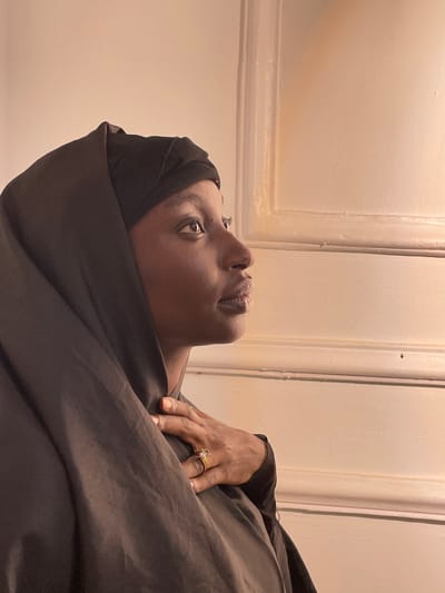 A young adult female, located in Barakin Akawo, Nigeria, is depicted in a profile portrait, facing right. She has a dark complexion and wears a black hijab draped over a visible black underscarf, along with a dark, loose-fitting garment. Her right hand is placed flat against her upper chest. A gold-toned ring, featuring a clear, square-cut stone, is visible on her right ring finger. Her gaze is directed upwards and to the right. The background consists of an off-white or cream-colored wall with a prominent vertical architectural molding, illuminated by soft, warm light.