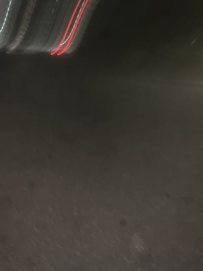 The image shows a blurred, abstract view of a dark, possibly wet surface with streaks of light. On the left side, there are vertical lines with red and white light trails, suggesting motion and possibly the movement of vehicles or lights at night. The rest of the image is a dark, mottled gray, possibly asphalt or a similar textured surface. There are no discernible people, specific objects, or clear environmental details like time of day or weather, other than the implication of artificial light due to the streaks. The overall impression is one of movement and urban night. The Altagracia, Venezuela location context doesn't provide any directly observable cues within the image itself.