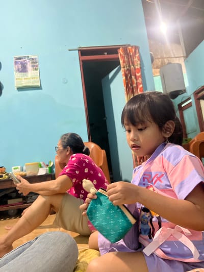 A young child wearing a pink and purple striped shirt and shorts is focused on a small teal pouch with a patterned design. The child is looking down, with their mouth slightly open, seemingly engaged in an activity with the pouch. Behind the child, another person, an adult woman wearing glasses and a pink shirt with a white circular pattern, is leaning forward and also appears to be interacting with something. The adult's hair is pulled back into a bun.
The scene takes place indoors, with light blue walls visible. A patterned curtain hangs in a doorway to the left. A calendar with images is attached to the wall on the left side of the frame, and there is a bright light source emanating from the ceiling on the upper left. The overall impression is of a casual, domestic setting. The image appears to be taken in the daytime, given the lighting. There is no information about the weather or specific emotions conveyed beyond the child's focused expression. No discernible text is visible within the image, other than potentially on the calendar, which is not clear enough to read. The overall orientation of the image is rotated. The location is Kecamatan Pati, Indonesia.