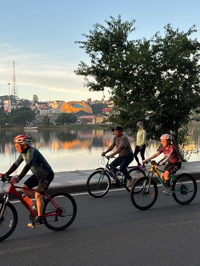 The image depicts a scenic view of Xuan Huong Lake in Da Lat, Vietnam, during a golden hour sunrise. In the foreground, four individuals are cycling along a paved path beside the lake. The first cyclist from the left is wearing a teal long-sleeved cycling jersey and black cycling shorts, with a red and white helmet. The second cyclist is a man in a grey t-shirt, dark pants, and a dark baseball cap, riding a blue bicycle. Behind him, a person in a light yellow hoodie and black pants is walking. The fourth cyclist, on the right, is a woman wearing a burgundy long-sleeved top and black cycling shorts, with a pink helmet.
The lake occupies the mid-ground, reflecting the warm hues of the sunrise and the surrounding cityscape. Across the lake, the Da Lat city skyline is visible, featuring a distinctive golden-domed building, residential houses with red roofs, and a tall radio tower on the left. The sky is a clear blue with a few wispy clouds, and the overall atmosphere suggests a peaceful morning activity. The lush green foliage of a large tree is prominent on the right side of the image, framing the scene.