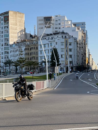 This image depicts an urban street scene in Tangier, Morocco, under a clear blue sky with distinct shadows indicating late afternoon. The foreground features a multi-lane asphalt road with white lane markings and directional arrows, including a central reservation. A fully-geared individual, wearing a black helmet and clothing, operates a black touring motorcycle equipped with saddlebags and gold-rimmed wheels, positioned centrally and moving from right to left across the frame.
To the right of the motorcycle, a paved pedestrian sidewalk runs parallel to the road. Two male pedestrians are observed walking away from the viewer; the leading individual wears dark clothing and a white cap, while the trailing one wears a light blue top. Further down the road on the right, a person rides a white scooter.
A white metal barrier, partially marked with red and white stripes, separates the roadway from a grassy median strip on the left, which contains several palm trees and cone-shaped evergreen trees. Modern, white, curved streetlights are spaced along the road and median.
The background is dominated by a dense arrangement of multi-story urban buildings. Most buildings are white or light-colored, featuring various architectural styles, balconies, and numerous windows. A prominent white building in the mid-ground displays the vertical blue sign "HOTEL MINARA." Rooftops throughout the cityscape are visible, showing fixtures such as satellite dishes, solar water heaters, and a cellular antenna on one building. A tall, unpainted building stands out on the far left.