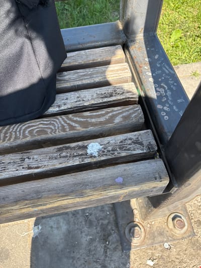 An outdoor shot of a section of a weathered wooden bench. The bench is constructed of several thick wooden planks supported by a dark gray metal frame. The wood shows signs of age and wear, with cracks and peeling paint. There are several splotches of white and purple residue on the wooden planks, possibly bird droppings or paint. A black bag is visible on the left side of the bench, partially obscuring the planks. The background consists of green grass and a concrete surface. The scene appears to be in a public outdoor space.