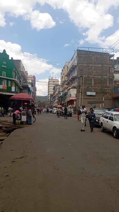 The image captures a daytime scene on Mburu Gichua Street in Nakuru, Kenya, oriented towards a distant hilly horizon. The sky overhead is partly cloudy with visible blue patches. The street itself is paved, exhibiting signs of wear, cracks, and uneven surfaces, particularly in the left foreground.

Multi-story commercial buildings line both sides of the street. On the left, a prominent green-painted building displays "1942 BUILDING" on its upper facade. Below it, a street vending operation is active, featuring a large red umbrella, a smaller pink umbrella, and a white vendor cart positioned on the uneven sidewalk.

Numerous individuals are present: pedestrians walk along both sides of the street and within the roadway. In the mid-ground on the right, a male subject in a light-colored collared shirt and dark trousers walks towards the viewer, holding a white object. A female subject in a dark dress walks slightly behind and to his left.

Vehicles include multiple parked cars along the right side of the street, one being a white station wagon. Several motorcycles (boda-bodas) are also visible, some parked and some with riders. Overhead power and utility lines crisscross the upper portion of the scene, connecting to poles and buildings. Buildings on the right side display various commercial signage, including a partially visible "HARDWARE" sign and a "MAGNAT PROPERTY" sign.