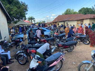 This image captures a bustling outdoor scene in Mararaba, Nigeria, characterized by a large gathering of people, numerous motorcycles, and several simple structures.

The foreground and midground are dominated by an unpaved, reddish-brown dirt ground, upon which approximately 20-30 motorcycles are parked. These motorcycles are primarily standard commuter models, featuring designs with chrome fenders, visible engines, and a mix of red, black, and silver color schemes. Some motorcycles, particularly those in the lower right, exhibit new plastic wrapping on their seats and handlebars. One light blue SUV is partially visible in the middle-left, parked among the motorcycles and people.

Numerous individuals, numbering between 50 and 70, are present across the scene. They are predominantly adults, dressed in a variety of traditional and casual attire, including colorful patterned dresses and wraps for women, often with head coverings, and tunics, shirts, and trousers for men, some with caps. People are observed standing in groups, conversing, and moving throughout the area. In the lower left, several men are seated on low stools, one in a white kaftan is looking at a mobile phone. In the center-left, a young man in a white t-shirt and green pants leans on a motorcycle. Towards the center-right, a woman is holding a large, round, flat object, possibly a tray or lid. Various individuals are either standing beside or sitting on parked motorcycles.

In the background, several single-story buildings with weathered, reddish-brown corrugated iron roofs and light-colored, possibly mud or concrete, walls are visible. These structures appear functional and simple. In the upper-left, two light-colored canopy tents are erected, under which a concentration of people suggests a social gathering or event. Large green trees, including a dense-leafed tree in the upper-left and palm trees in the mid-background, provide natural elements. The sky above is clear to partly cloudy blue, with visible power lines traversing the upper portion of the frame.