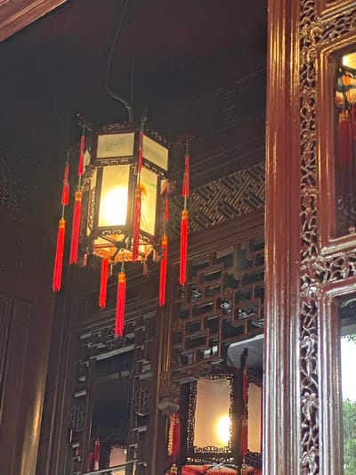 The image captures an ornate, traditional Chinese lantern hanging indoors, likely in Shanghai, China, given the architectural style. The lantern is hexagonal with illuminated panels and adorned with numerous red tassels. It hangs from the dark wooden ceiling of a room featuring intricate wooden lattice work and rich, dark wood paneling. The warm glow of the lantern illuminates the scene, casting soft light on the decorative elements. To the right, a richly carved wooden pillar with ornate detailing frames part of the view. The overall impression is one of traditional elegance and cultural richness.