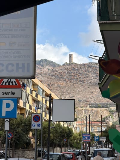 This image, captured in Maddaloni, Italy, presents a lively street scene beneath a clear blue sky with some scattered clouds. The street is lined with multi-story buildings, characterized by balconies and a mix of residential and commercial fronts. Parked vehicles, including a red car and a white van, line the street, suggesting an urban environment.
In the background, a large, rocky mountain rises, sparsely covered with vegetation, and topped by a distinct, ancient-looking stone tower. This geographical feature provides a dramatic backdrop to the townscape. The bright daylight indicates a time between late morning and afternoon.
Numerous street signs are visible. There are multiple blue "P" signs indicating parking, one specifically noting "P&" for disabled parking. A "no parking" sign (red circle with a diagonal line) is also present, along with directional arrows. A street name sign reads "S. Gentile." A blue advertising banner prominently displays "DETERSFUSO Risparmio," likely for a detergent store. Another sign features the word "serie." On the far left, a large, partially obscured sign shows mirrored text, including fragments like "LEBIV CVDCEL" and "CCHI."
A man with light hair is partially visible on the right, near a white vehicle, suggesting a driver or pedestrian. The overall scene depicts typical daily life in an Italian town nestled against a mountainous landscape.