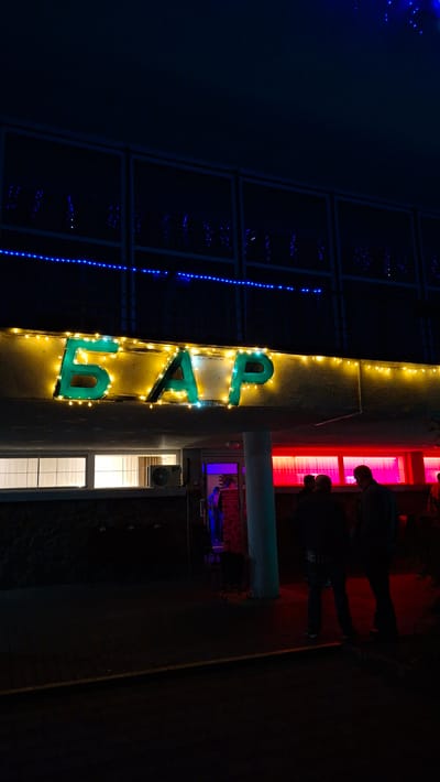 At night in Нарацкі сельскі Савет, Belarus, the vertically oriented image shows the illuminated sign for a "BAR" in bright green letters outlined with warm yellow lights. The sign is attached to a light-colored vertical wall. Blue lights are visible on a darker building to the left. Silhouettes of two people are seen near red-lit windows to the right. The foreground is dark.
