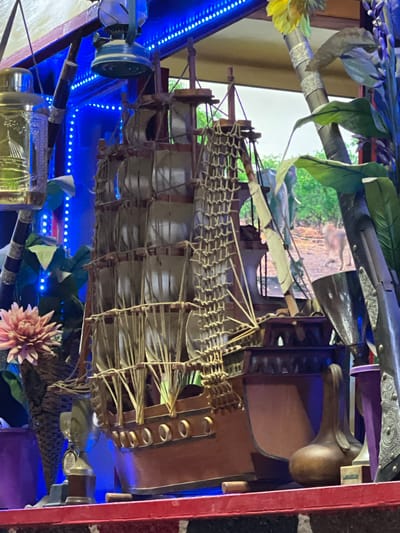 A sideways close-up photograph reveals a maritime display encased within what appears to be a cabinet or display case. At the core of the image is a miniature ship, meticulously detailed with sails and rigging. The ship is surrounded by various objects, including bottles and decorative flowers. A gun is visible along the top. The scene is illuminated by vibrant blue LED lights. The background includes what appears to be a window offering a glimpse of greenery. The overall composition suggests a curated collection of maritime-themed items. Considering the unusual arrangement and collection of items, it is not possible to determine the exact location in Meknes, Morocco based on this single image.