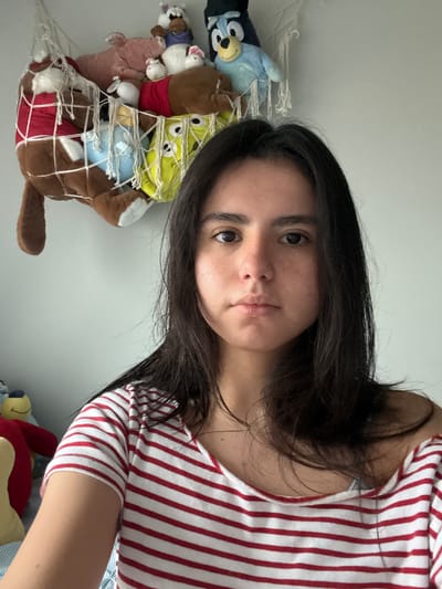 A young adult female is the main subject of the image, wearing a red and white striped shirt. Her dark hair is shoulder length. She is taking a selfie indoors, with a white wall as the background. A net filled with various stuffed animals is visible to the left. The location is not discernible from the image, so an association with the city of Austin is speculative.